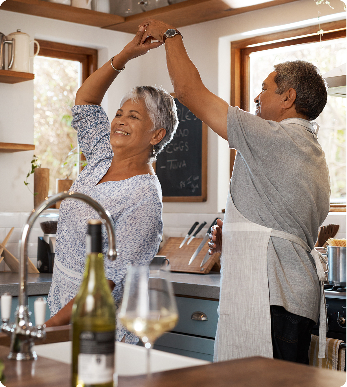Smiling senior couple dancing together in a cozy kitchen, wearing aprons, with wine and a glass on the counter in the foreground.